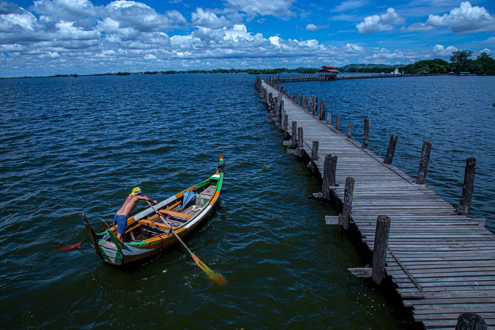 Man rowing a wooden boat beside the U Bein Bridge near Mandalay, the kind of scene a Burma river cruise passes